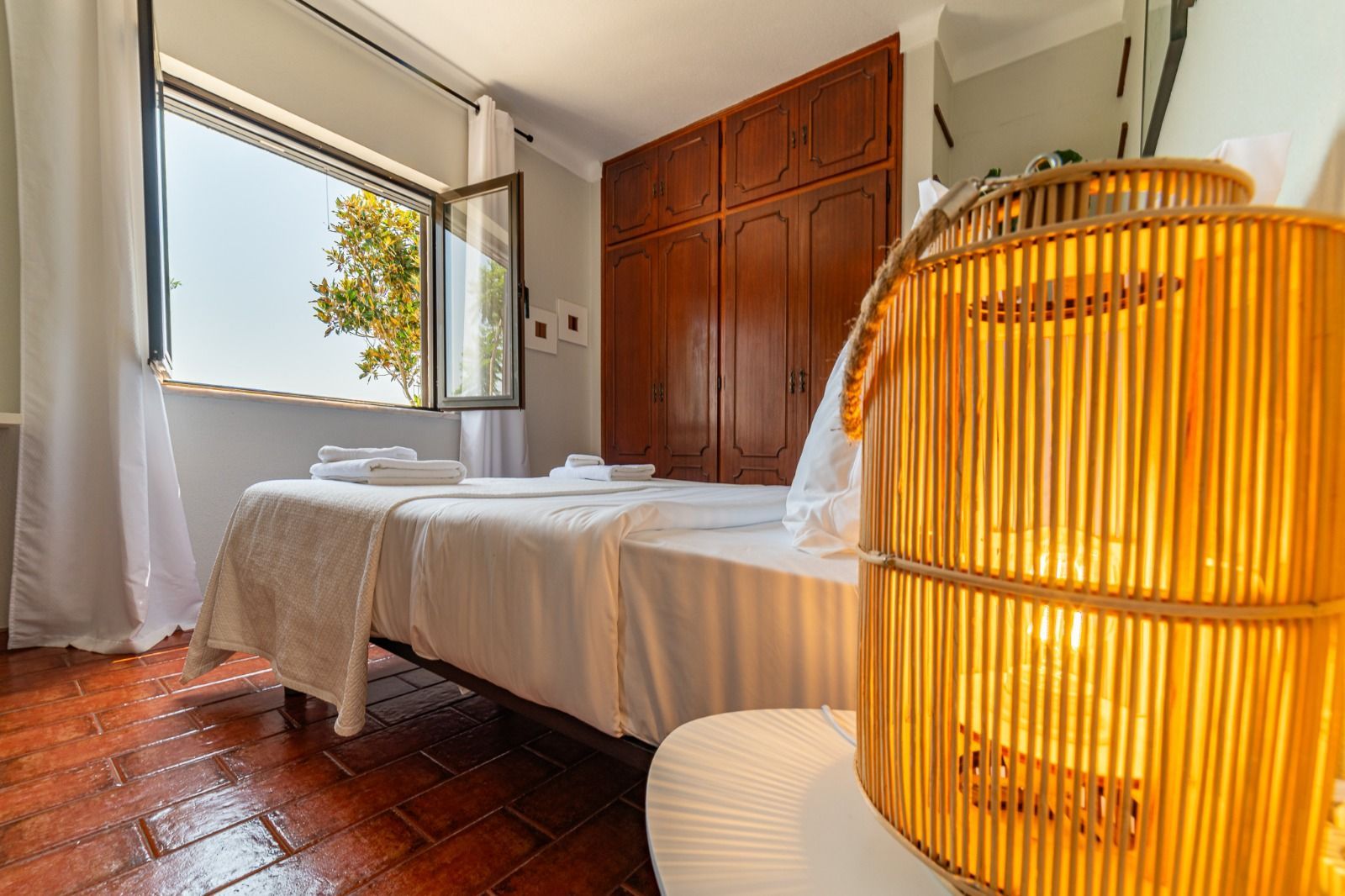 Bright bedroom with twin beds, open window, wooden wardrobe, and a woven lamp in the foreground