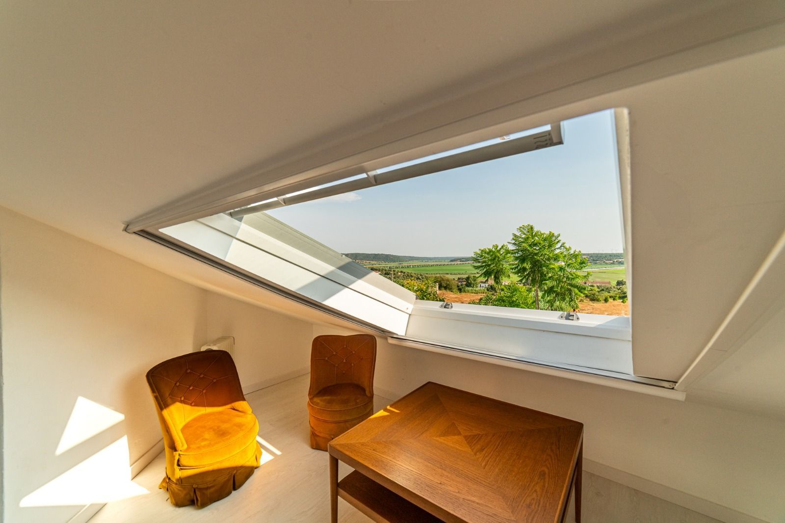Attic nook with slanted skylight, yellow chair, wooden table, and view of green landscape.