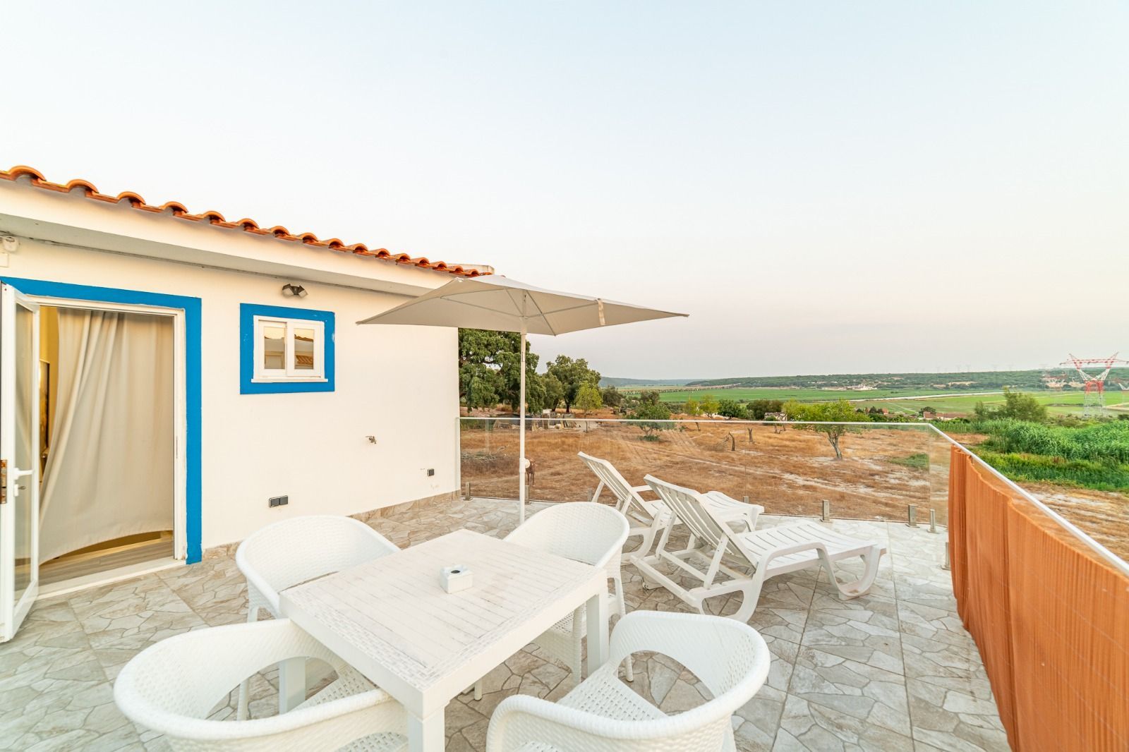 White patio with table and chairs beside a house, overlooking a green countryside view
