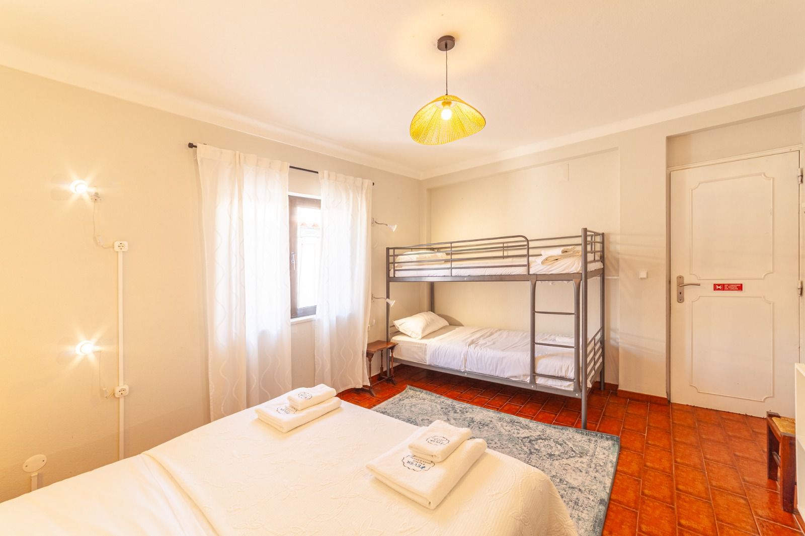 Bright bedroom with white bed, bunk bed, wooden floor, and sunlit windows