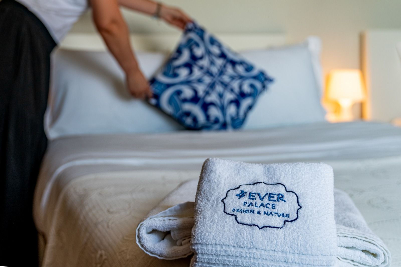 Hotel bed being made with white towels and a blue patterned pillow in the background
