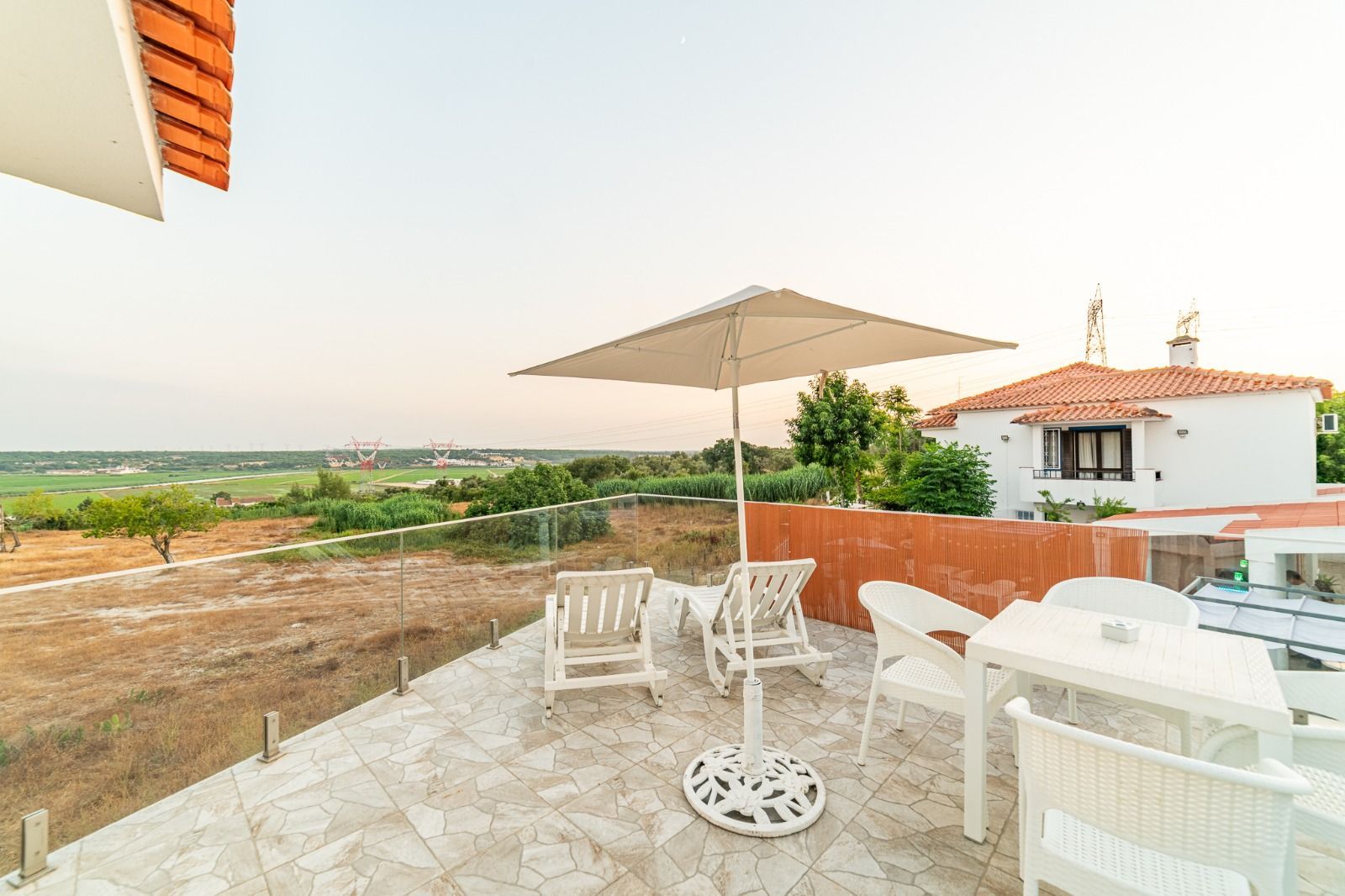 Patio with white table and chairs under umbrella, overlooking open fields and a small house at sunset