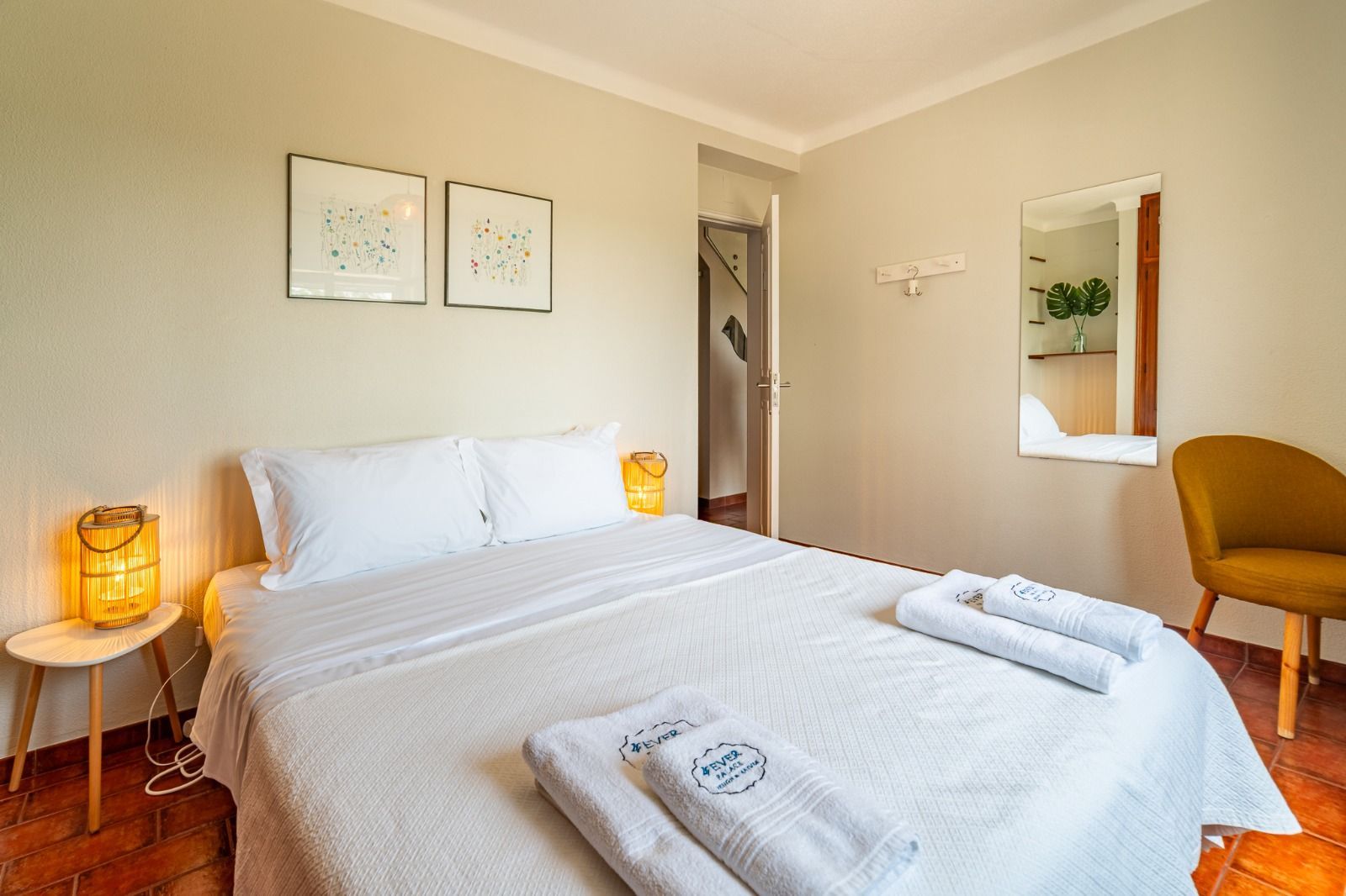 Bright bedroom with a neatly made bed, bedside lamps, wall art, and a chair by a mirror.
