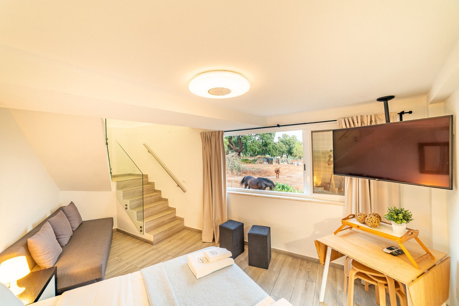 Bright living room with sofa, staircase, dining table, wall TV, and a large window