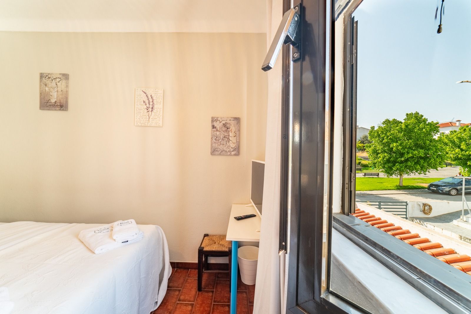 Bright bedroom with a neatly made white bed and an open window overlooking trees and a street.