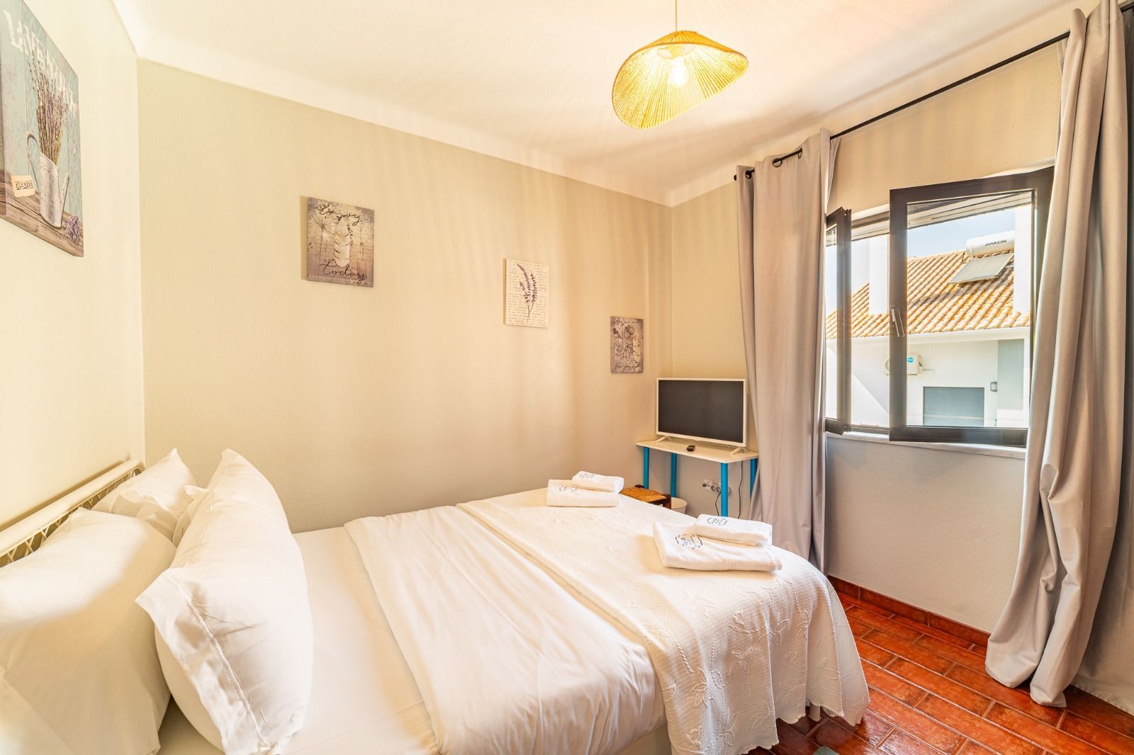 Bright bedroom with a neatly made bed, beige walls, and a window with gray curtains.