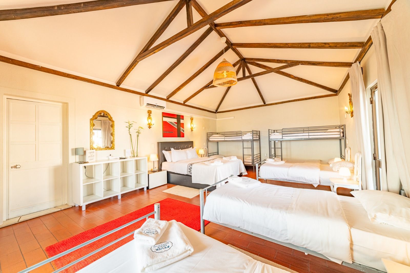 Bright airy bedroom with wooden beams, white walls, and two neatly made beds on a red floor.