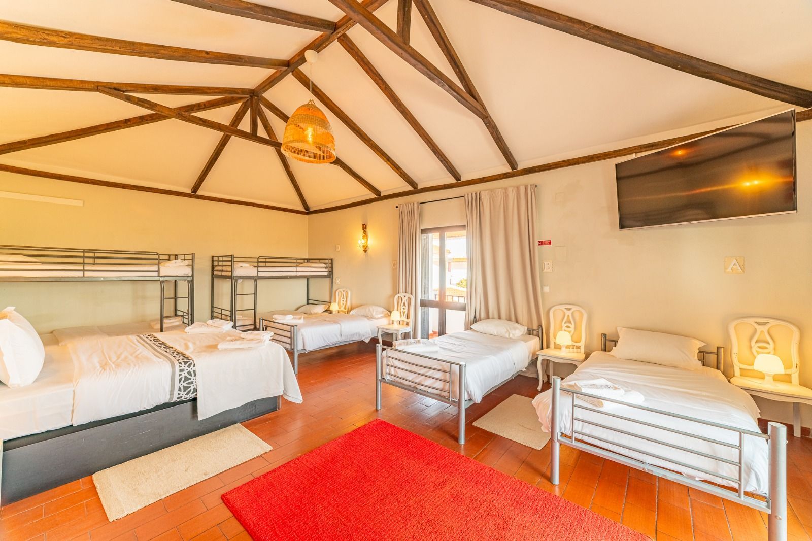 Bright bedroom with four beds, red rug, exposed beams, and a wall-mounted TV