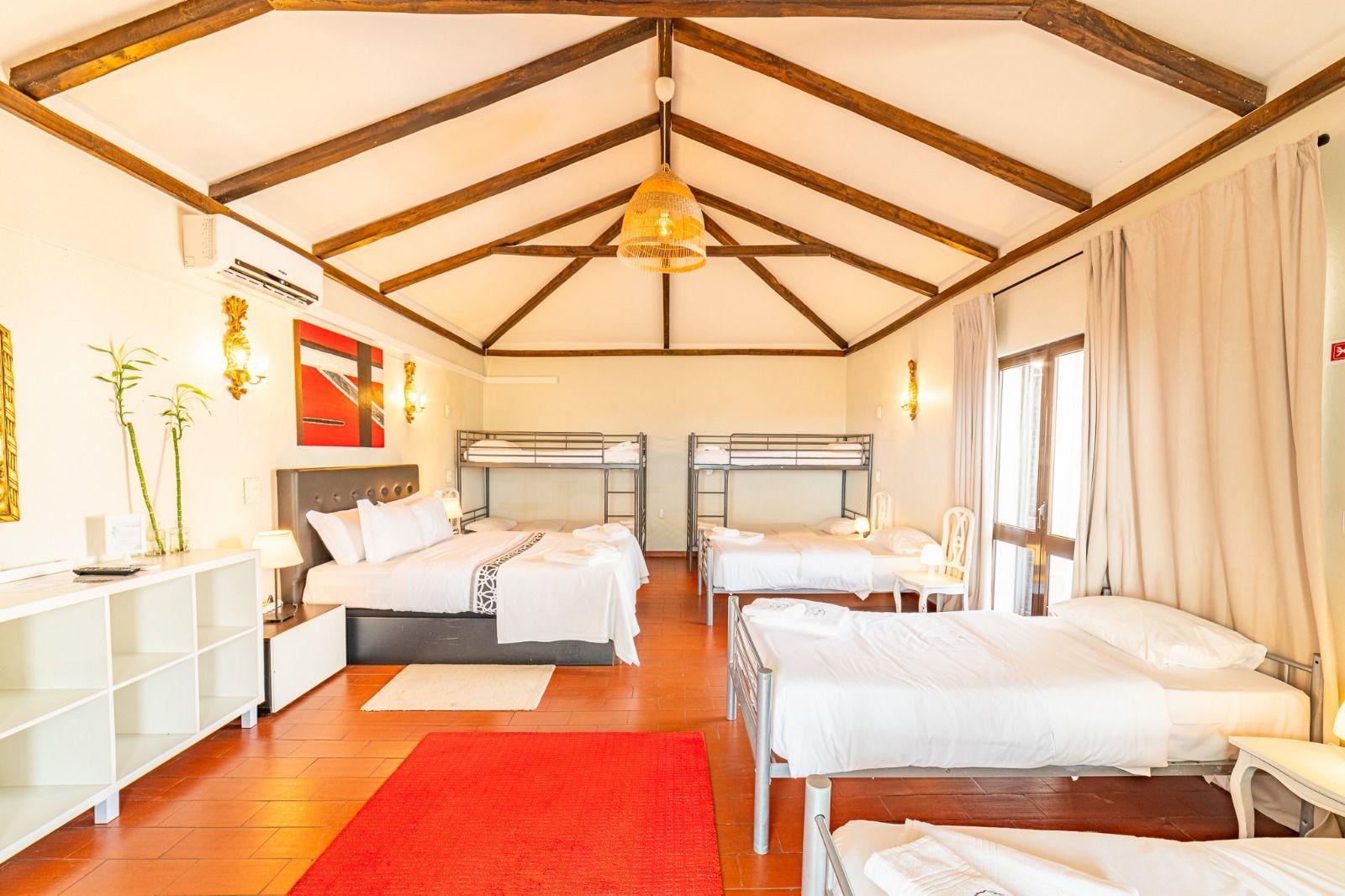 Bright bedroom with two beds, red rug, white curtains, and wooden ceiling beams