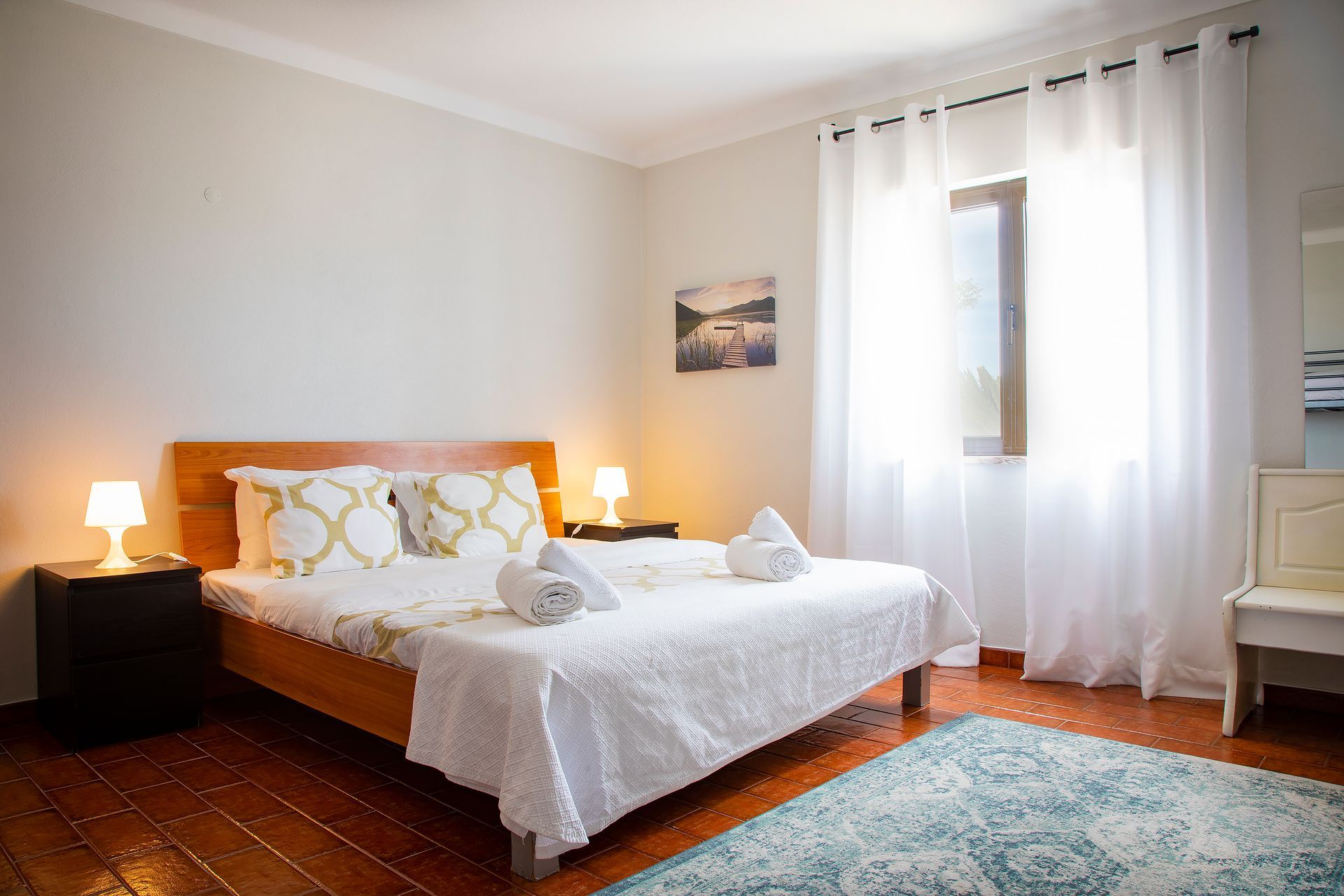 Bright bedroom with a wooden bed, white bedding, bedside lamps, and a window with sheer curtains.