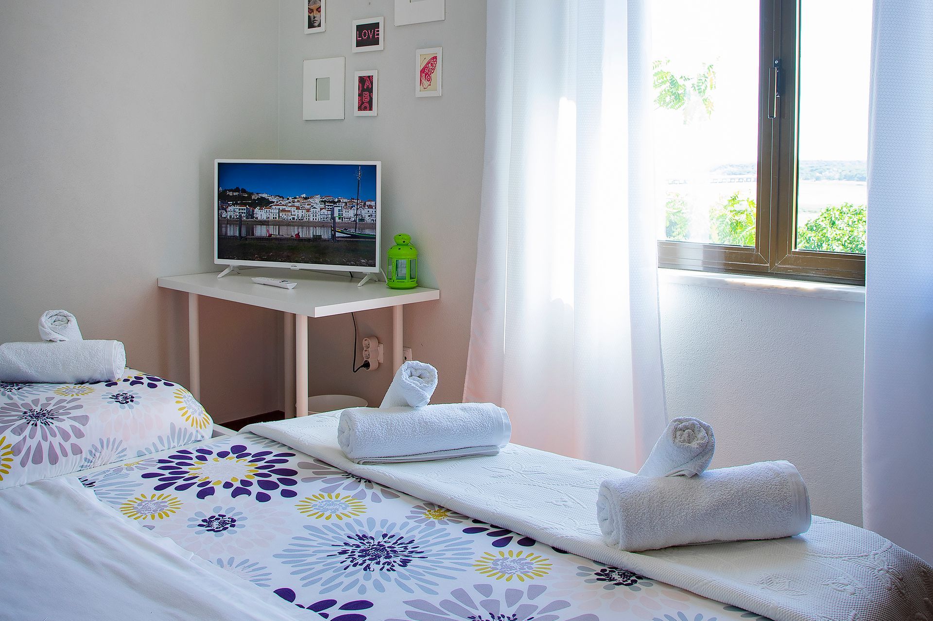 Bright bedroom with a bed, TV on a desk, and sunlight through a window with white curtains.