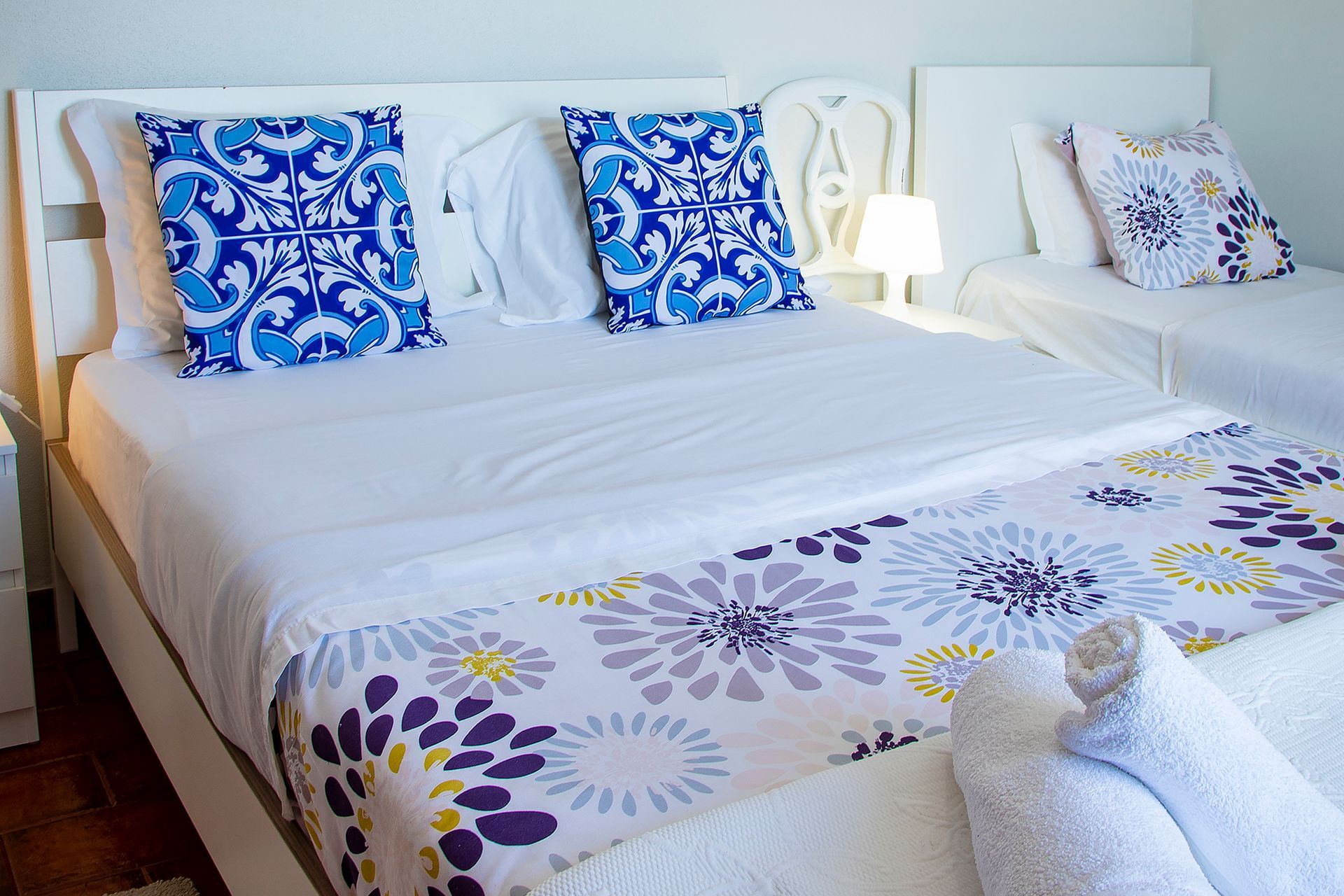 White bedroom with a neatly made bed, blue patterned pillows, and a floral bedspread with a bedside lamp.