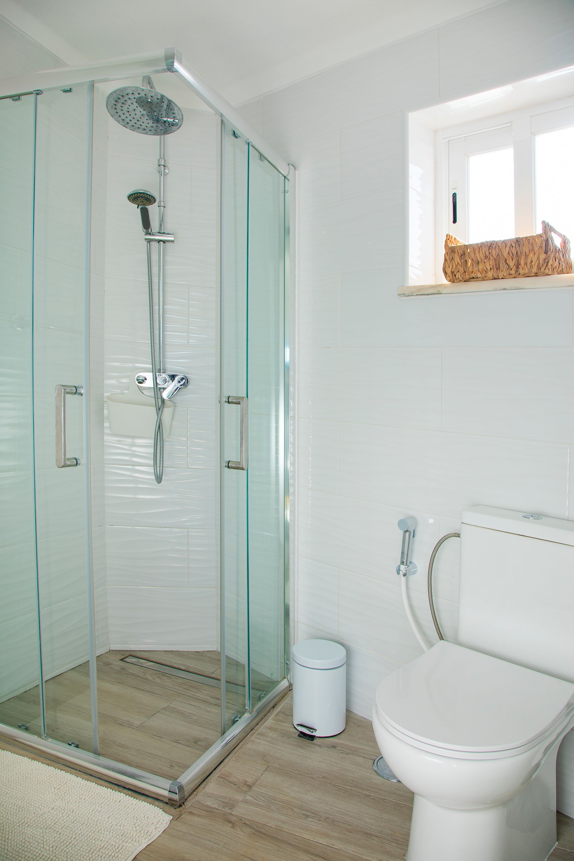 Small modern bathroom with glass shower, white toilet, and window with wicker basket shelf.