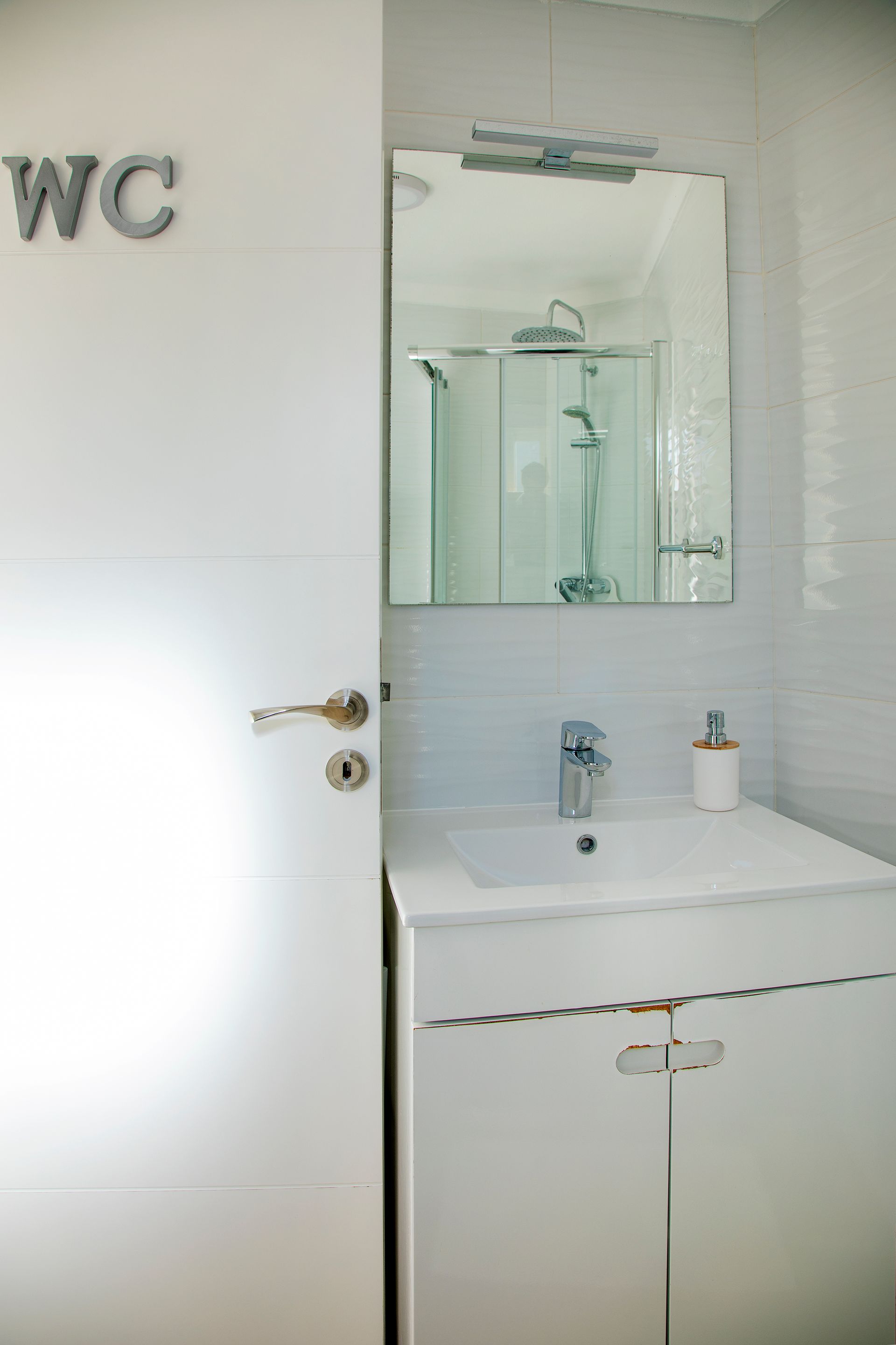 Small white bathroom sink with mirror and cabinet, next to a door marked “WC”.