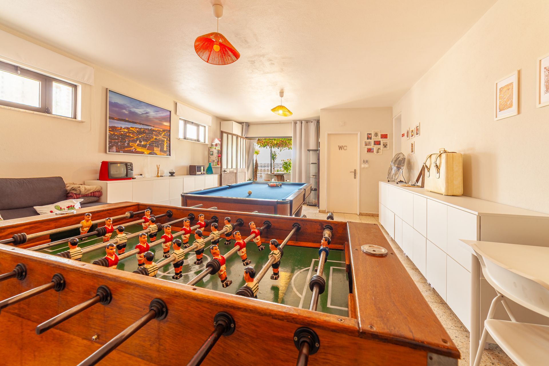 Bright game room with foosball and pool tables, white cabinets, wall art, and hanging lamps