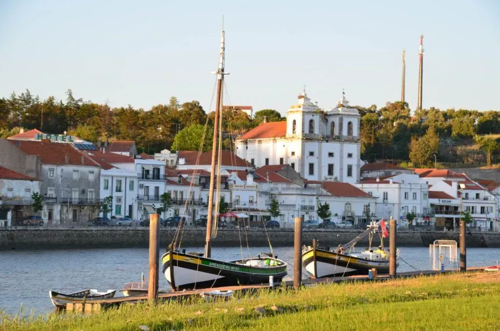 Riverfront village with white houses, a docked boat, and green grassy foreground at sunset