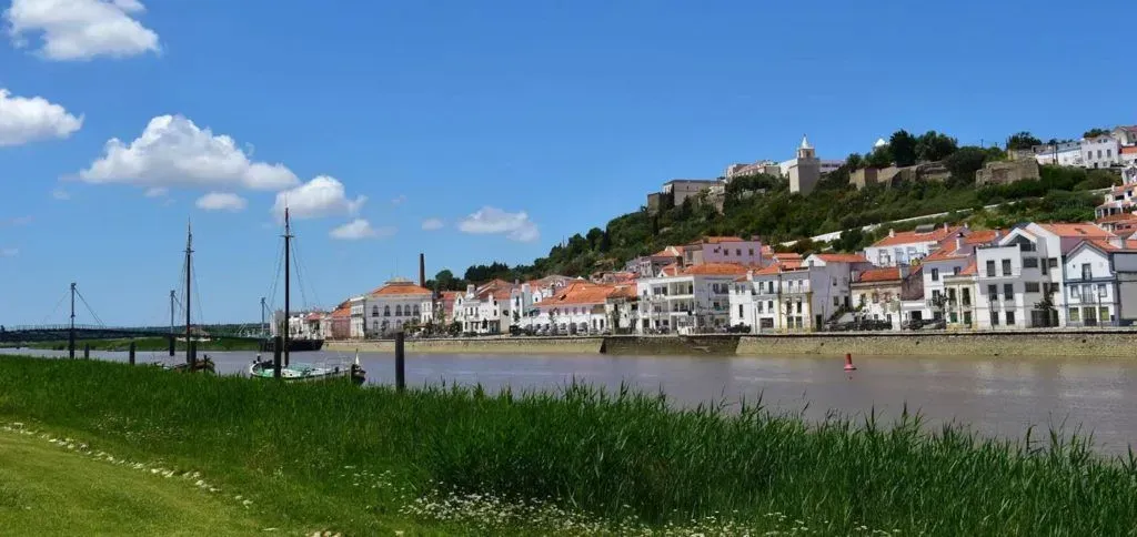 Riverside town with white houses and red roofs under a blue sky, beside a grassy bank and calm water