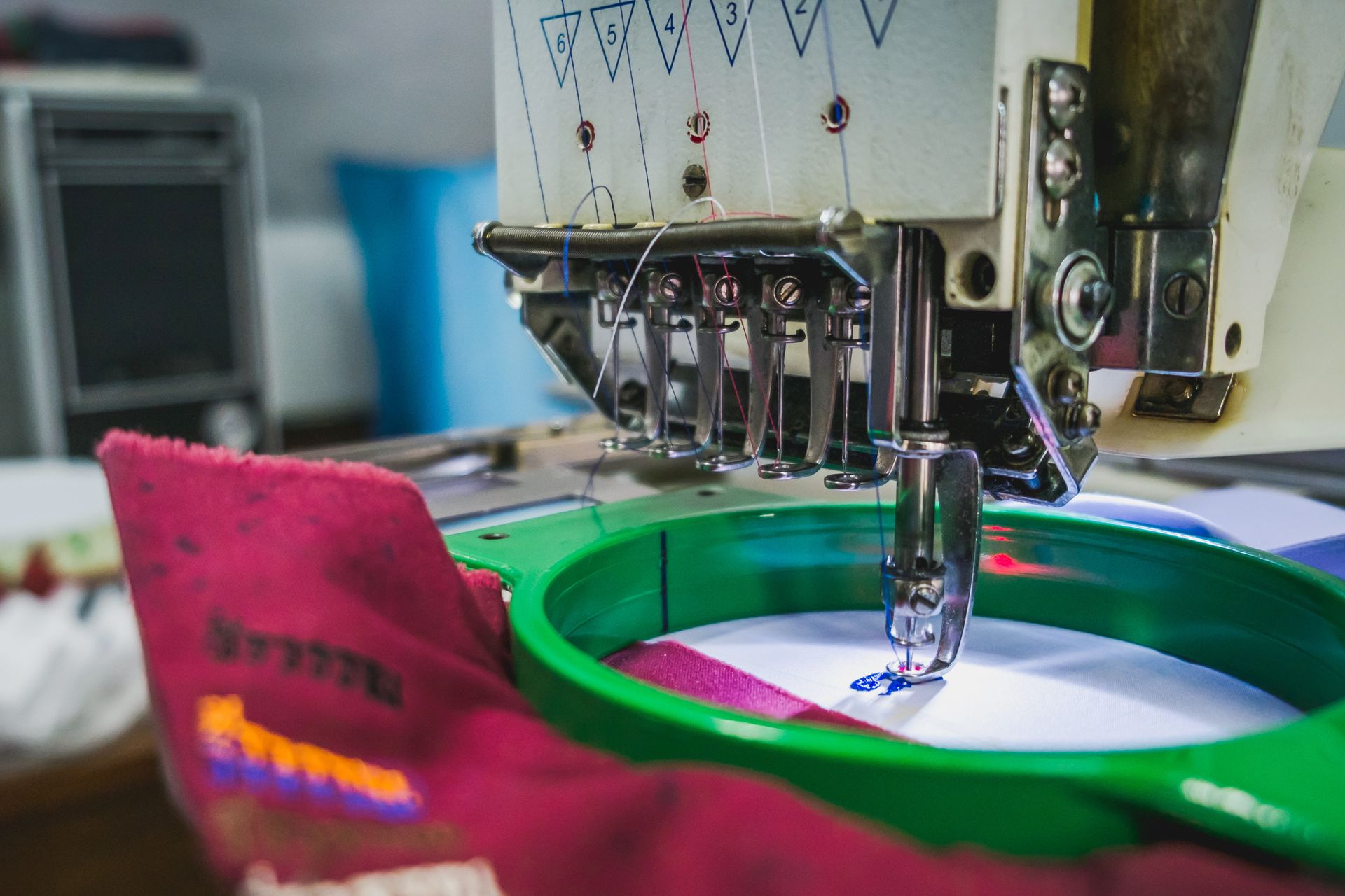 Una máquina de bordar industrial cosiendo un patrón sobre una tela roja sostenida en un aro de color verde brillante.