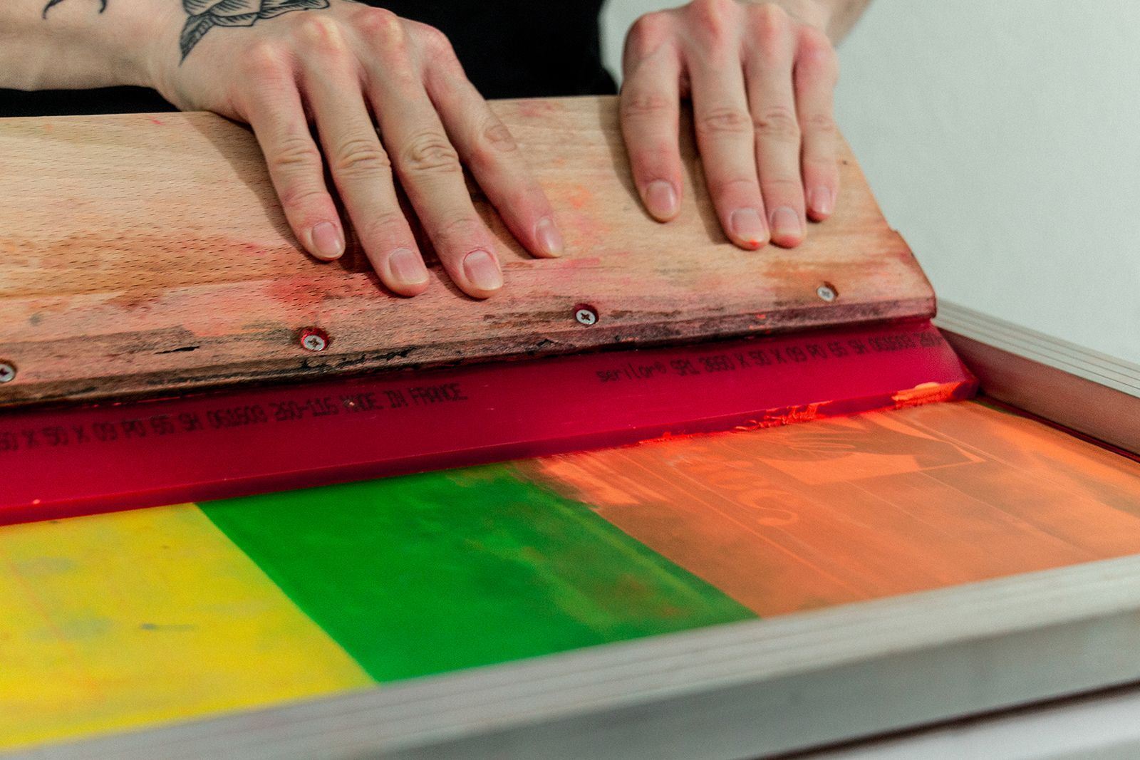 Unas manos presionan una espátula sobre un marco de serigrafía con tiras de tinta amarilla, verde y naranja.