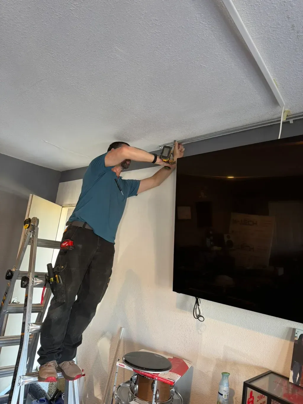 Person using a drill to install something near the ceiling in a living room beside a wall-mounted TV.