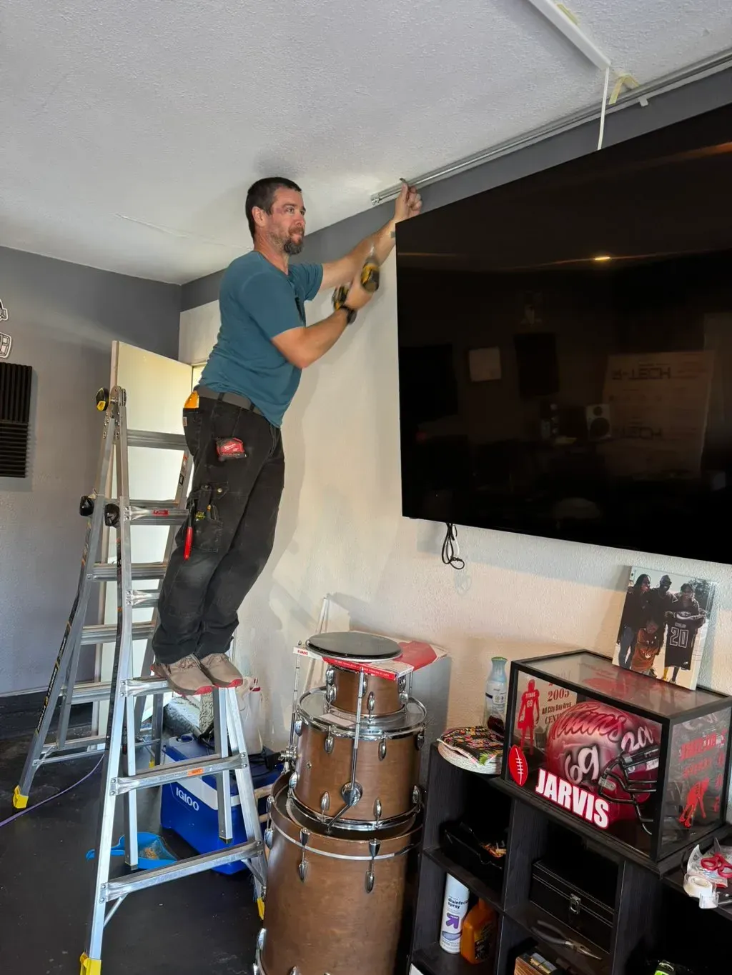 Man standing on a ladder painting the upper wall near a large TV in a room under renovation