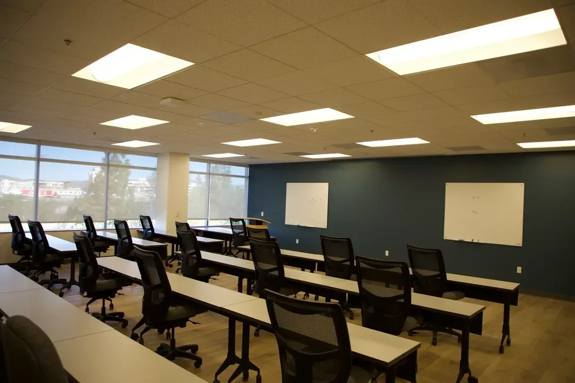 Empty classroom with rows of desks, chairs, and whiteboards under bright ceiling lights.