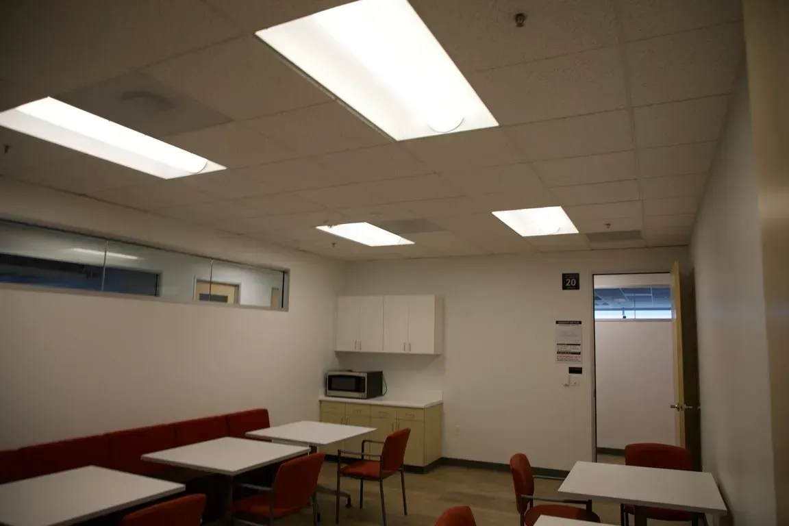 Empty break room with fluorescent lights, tables, red chairs, cabinets, and a white door.
