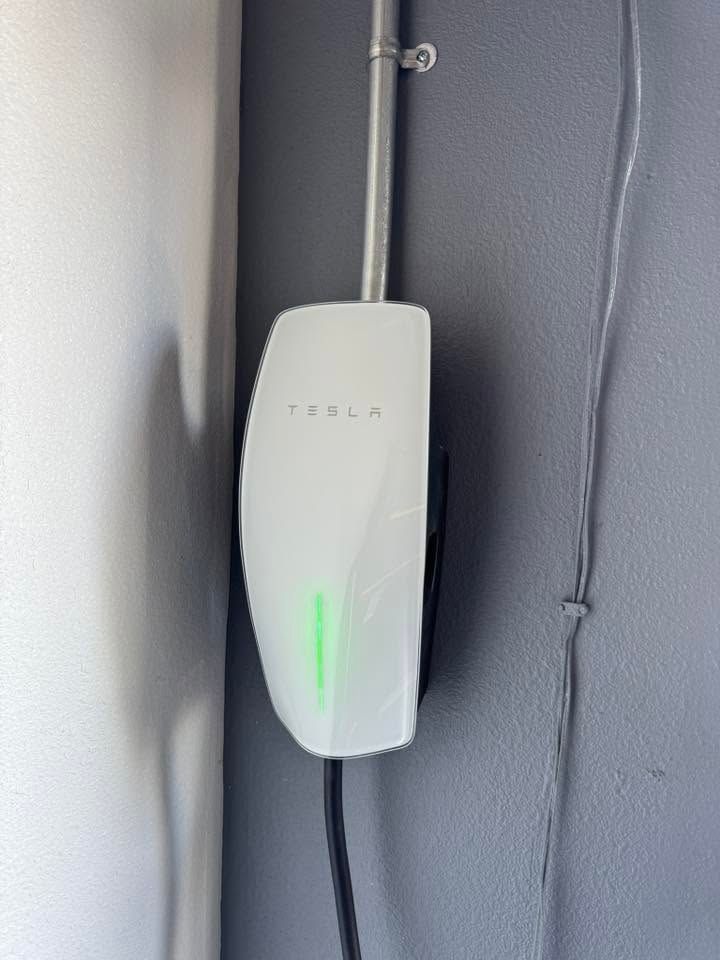 White wall-mounted device with a green light, attached to a gray wall by cables.