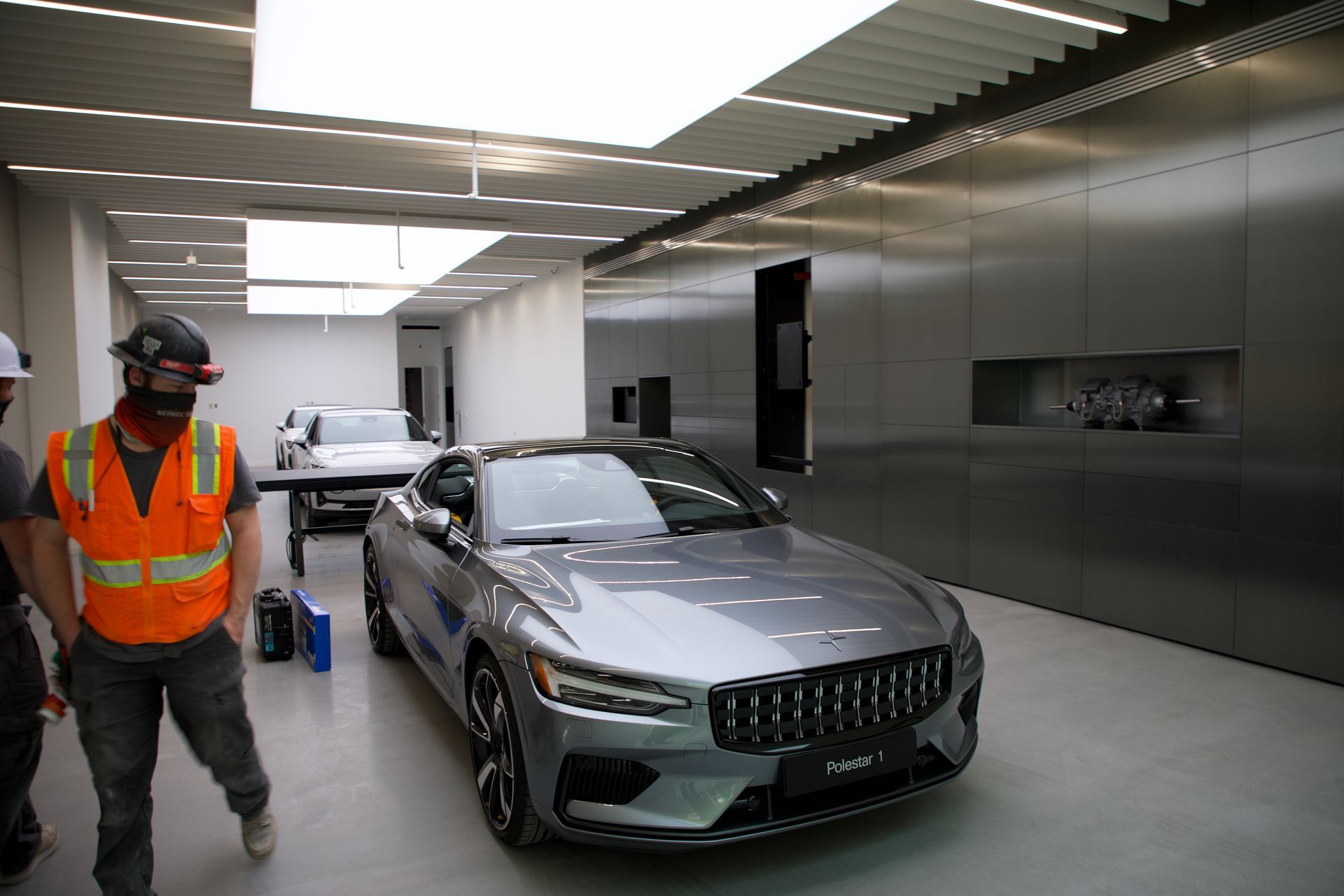 Worker in orange safety vest stands beside a gray sports car in a modern indoor garage.