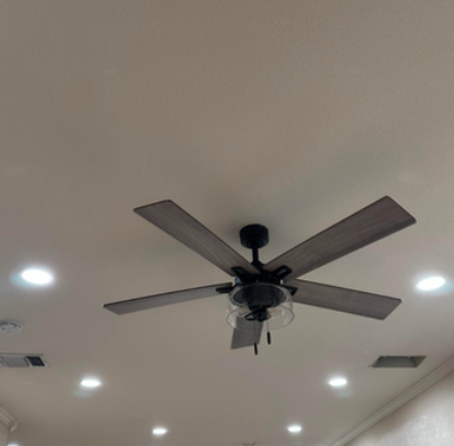 Ceiling fan with dark blades on a white ceiling with recessed lights and air vents.