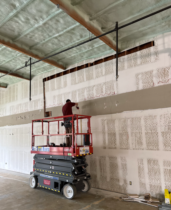 Worker on a red lift applying or inspecting wall panels inside an unfinished industrial building.
