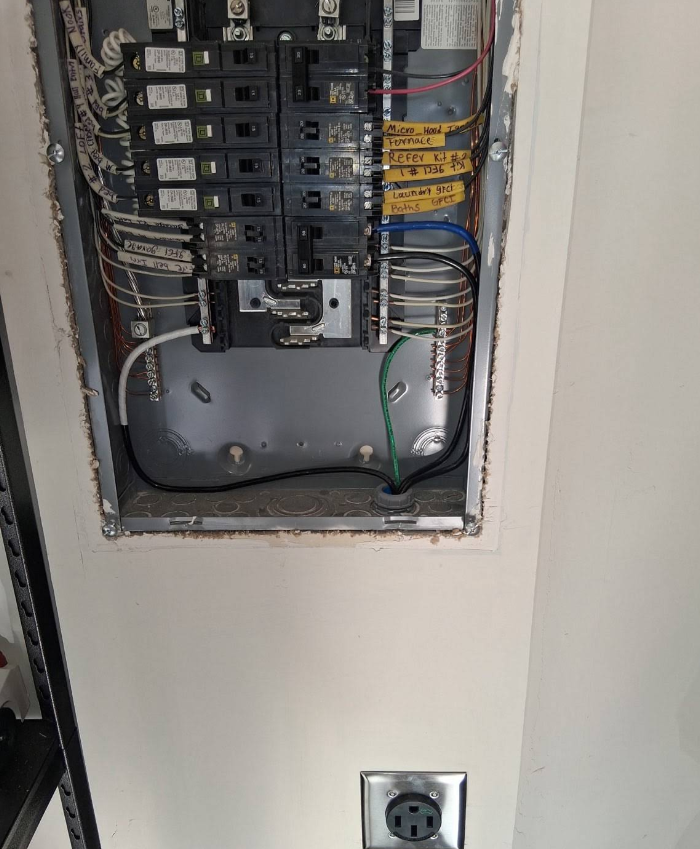 Open electrical breaker panel with wires exposed in a wall recess, outlet below.