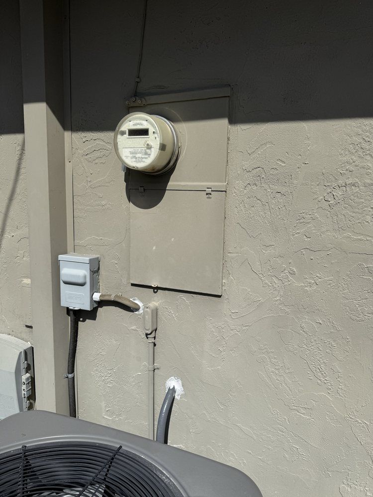 Electric meter and utility boxes mounted on a beige exterior wall beside piping and an air conditioner unit