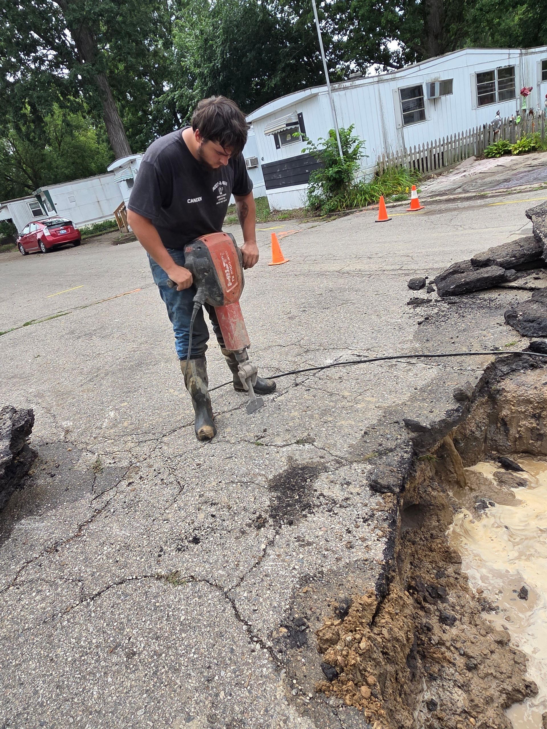 Worker using a jackhammer to break cracked pavement near a large sinkhole on a road.