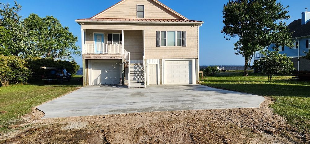 Two-story beige house with garage, balcony, and large concrete driveway in a sunny suburban setting