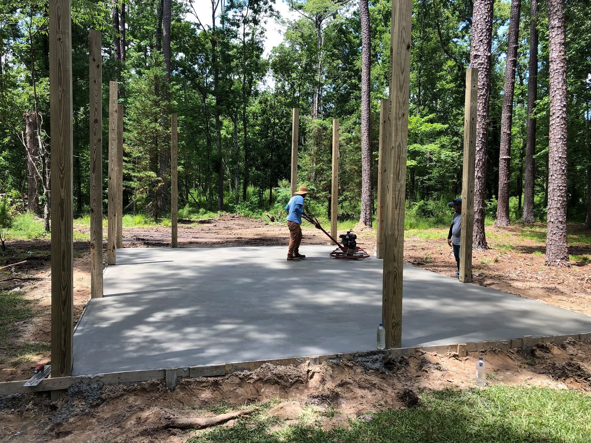 Person using a power trowel to smooth a fresh concrete slab in a wooded area.