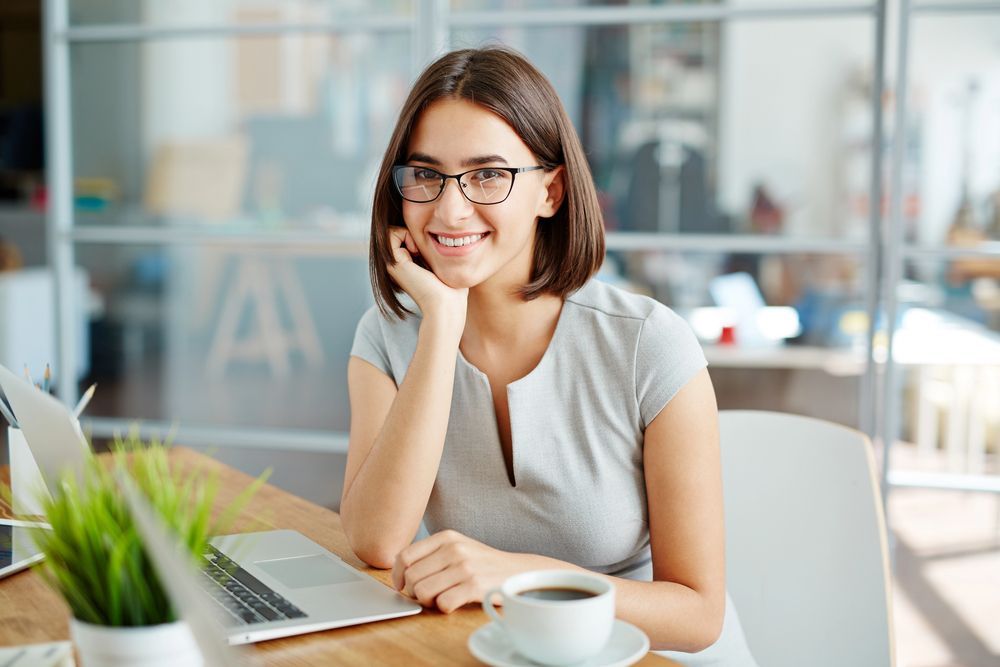 A woman is sitting at a desk with a laptop and a cup of coffee.