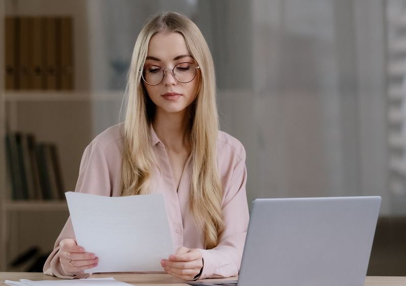 A woman is sitting at a desk with a laptop and holding a piece of paper.
