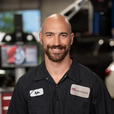 Bald, smiling man with a beard in a mechanic's uniform, in a garage setting. He has name badges and appears to be looking at the camera. | RX Automotive