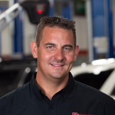 Man in a black shirt smiling at the camera, in an auto repair shop. | RX Automotive