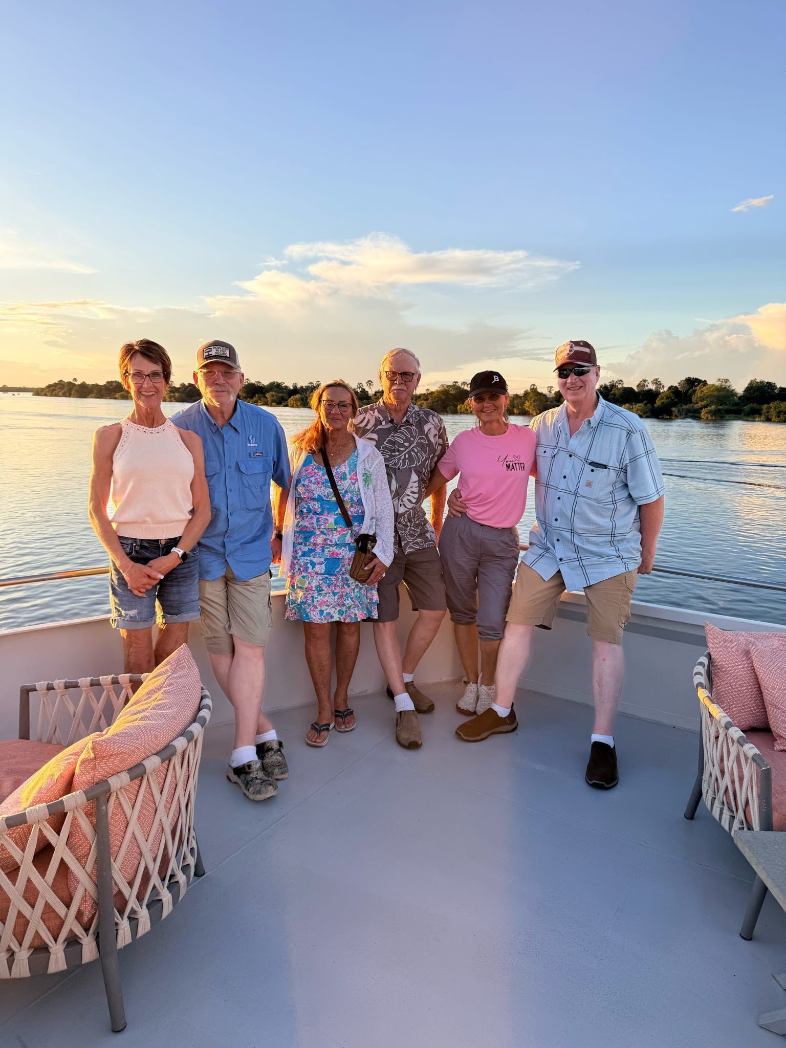 Group of five smiling friends walking on a boat deck at sunset by calm water