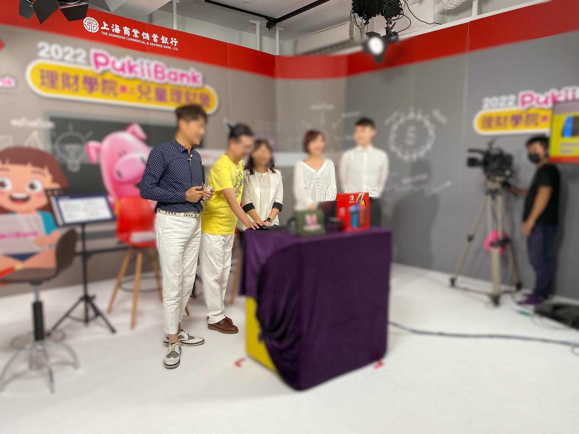 電視演播室裡的人們。主持人圍坐在桌子旁，桌上擺滿了禮物。舞台背景為紅色和黃色。