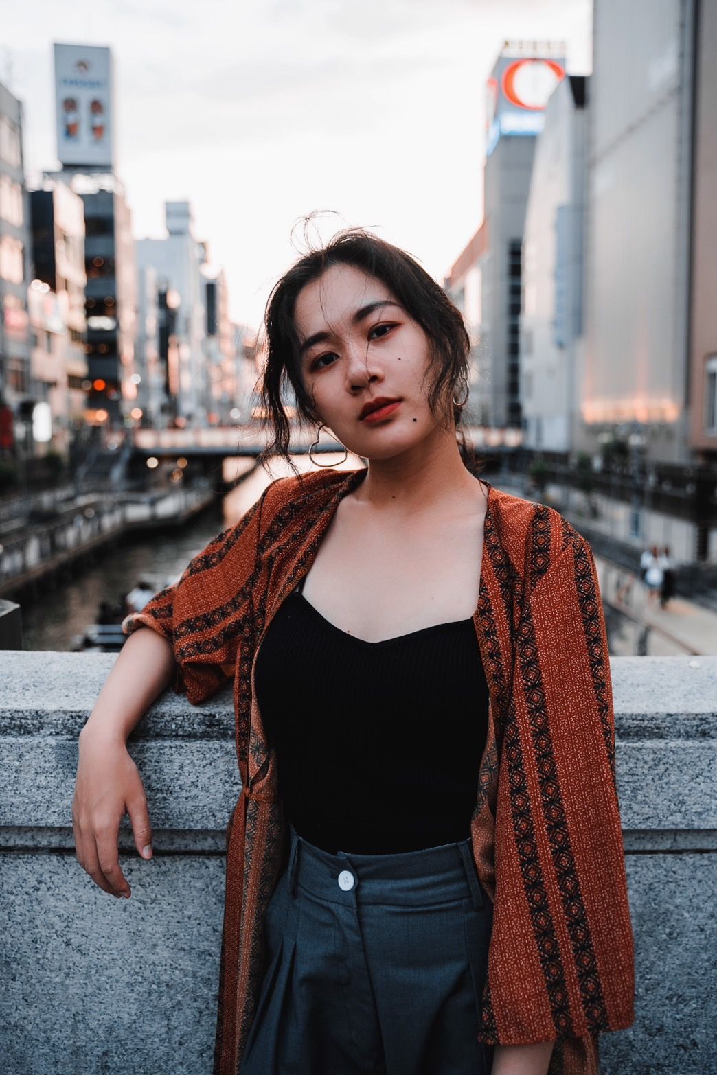 在城市環境中，一名女子倚靠在石障上，身穿黑色上衣和圖案的夾克。