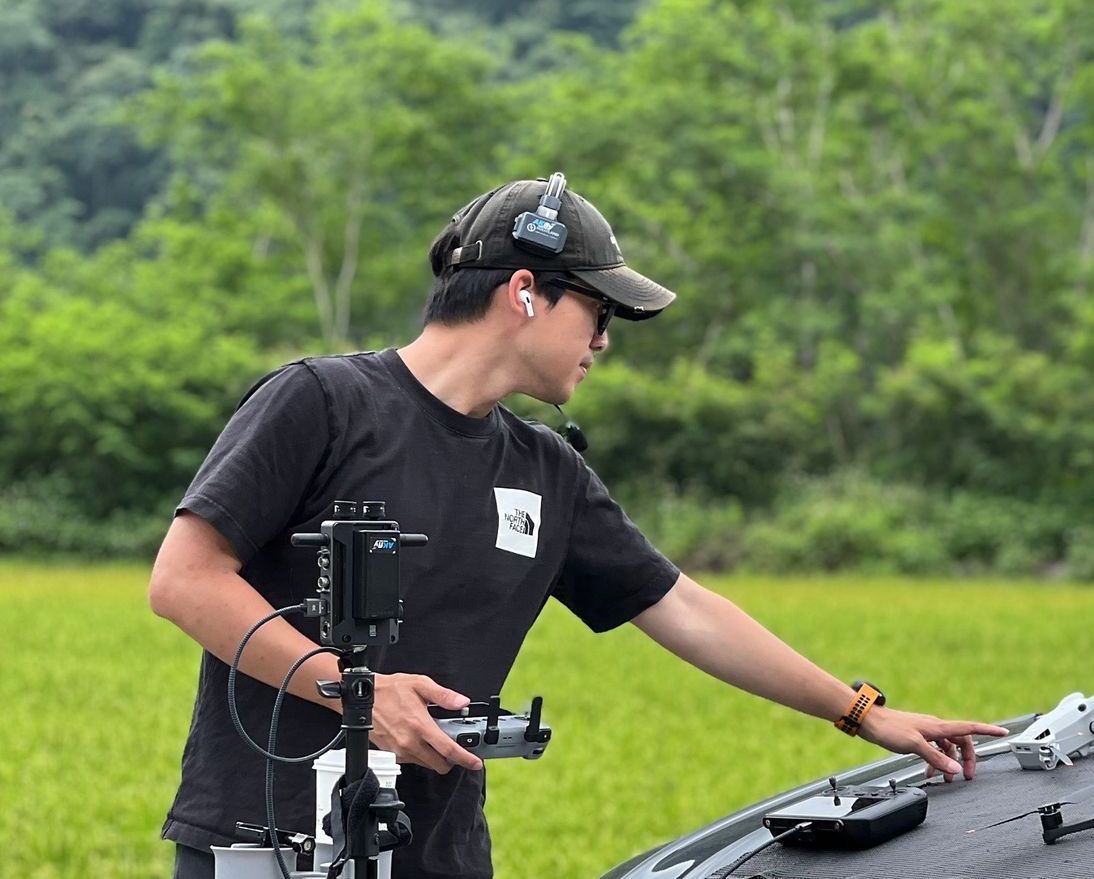 身穿黑色襯衫、頭戴黑色帽子的男子在戶外操作無人機，指著無人機表面。背景是綠色的田野和樹木。