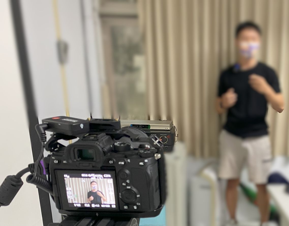 攝影機拍攝到一名身穿短褲和黑色襯衫的男子，他站在掛著米色窗簾的窗戶前做手勢。