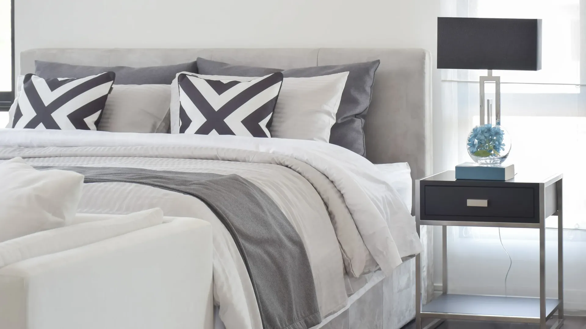 Modern bedroom with a grey upholstered bed, white and grey bedding, and a sleek nightstand with a lamp.