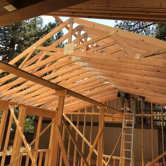 A wooden roof frame is under construction with a worker on a ladder, set against a background of trees.