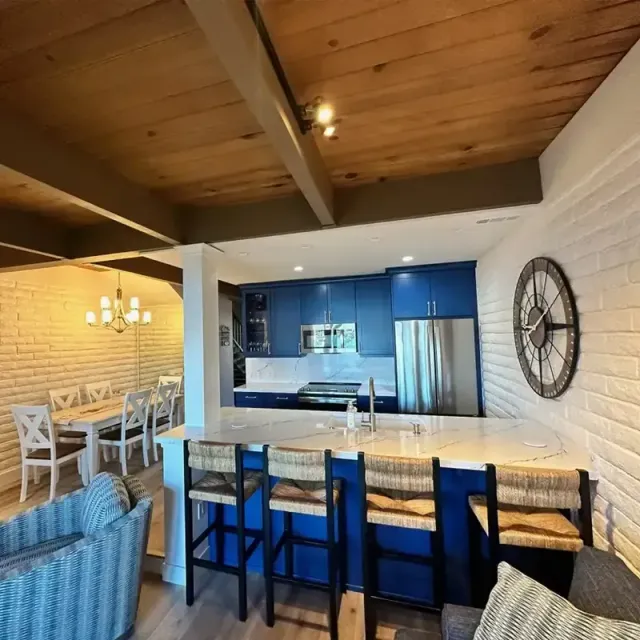 A kitchen with blue cabinets, white countertops, and bar seating, next to a dining area with a white table and brick walls.
