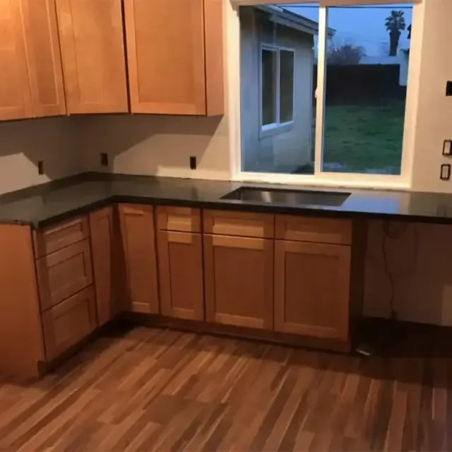 A kitchen with light wood cabinets, dark countertops, a stainless steel sink, and wood-patterned flooring.