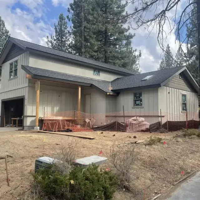 A modern two-story home under construction with light-colored siding, dark trim, and a roof, set in a wooded area.